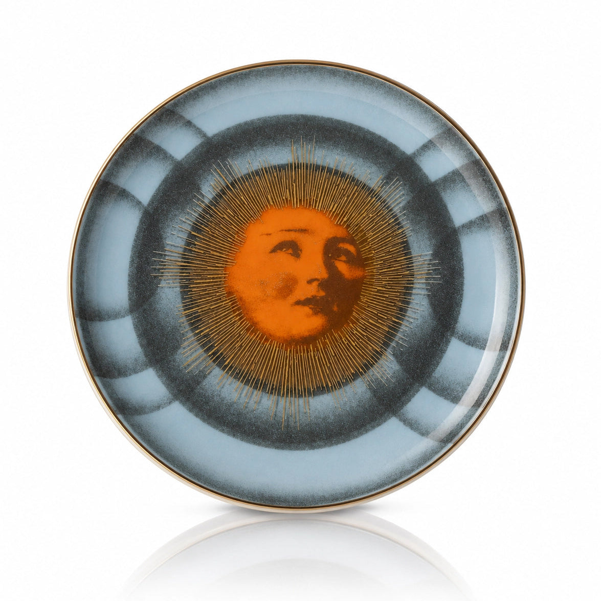 A round ceramic plate featuring a central sun motif with a stylized face and radiating gold lines, surrounded by concentric rings of dark gray and light blue, finished with a thin gold rim.