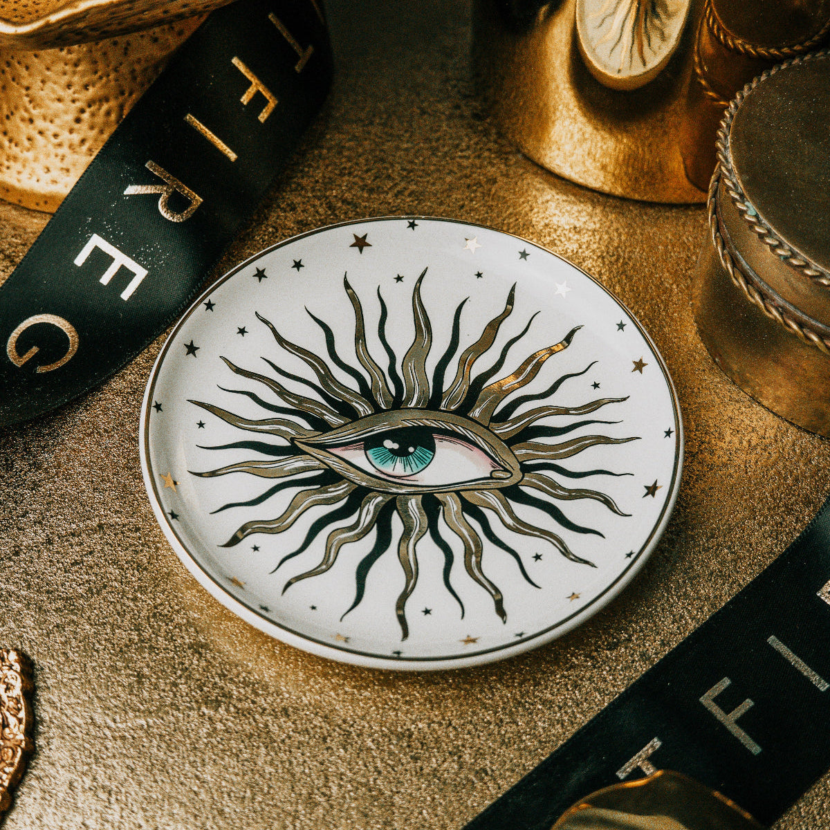 A round white ceramic plate featuring a detailed illustration of a single eye surrounded by radiating black and gold rays, styled on a textured gold surface with dark ribbons and metallic props.