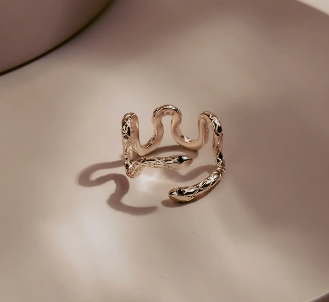 Sleek and hypnotic, the Serpent Coil Ring embodies power and transformation. Crafted in gleaming 14k gold plate over sterling silver, the coiled design wraps gracefully around the finger, ending in twin sculpted snake heads with dark alluring eyes. Adjustable for a perfect fit.