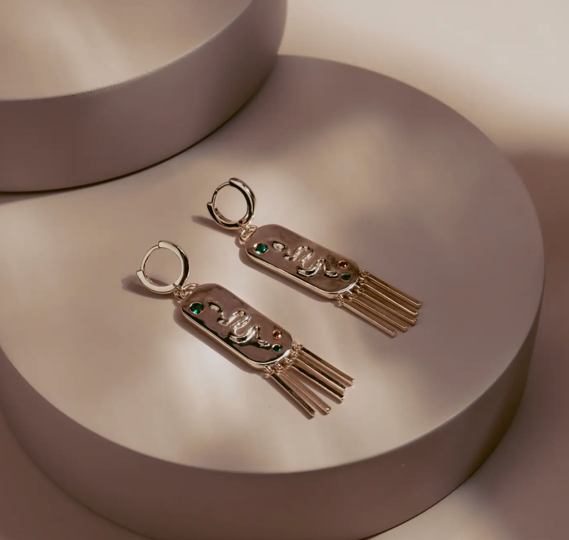 The Serpent Fringe Earrings fuse bold design with fluid movement. Crafted in 14k gold plated over silver, each piece features a raised serpent motif accented with deep green and red stones. The rectangular base flows into fine golden fringe, creating a striking play of texture and light. Confident and sculptural, they bring a touch of power and sophistication to any look.