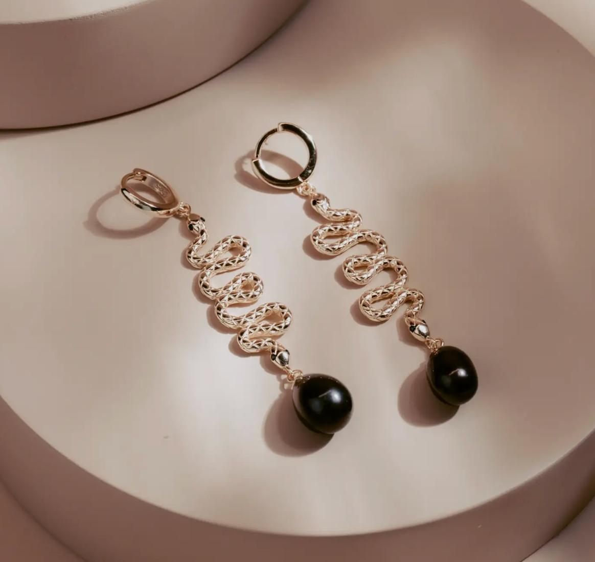 The Serpent Drop Earrings bring a sleek balance of movement and form. Crafted in 14k gold plated over silver, each earring features a sinuous snake design that flows into a dark teardrop pearl. Elegant and commanding, they capture light with every turn, embodying quiet power and refined allure.