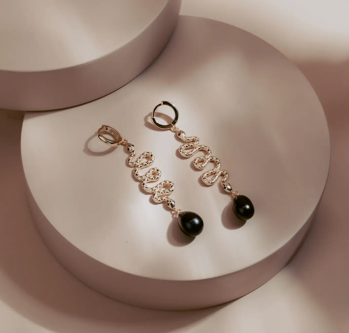 The Serpent Drop Earrings bring a sleek balance of movement and form. Crafted in 14k gold plated over silver, each earring features a sinuous snake design that flows into a dark teardrop pearl. Elegant and commanding, they capture light with every turn, embodying quiet power and refined allure.