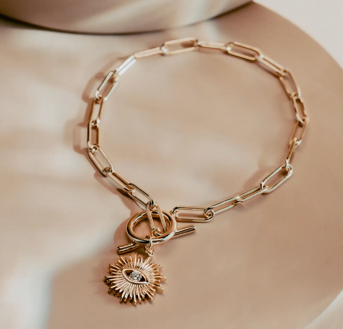 The Golden Eye Bracelet blends sleek design with symbolic detail. Crafted in 14k gold plated over silver, it features a bold paperclip chain and a front toggle clasp accented by a radiant sunburst charm set with a clear crystal eye. Polished and versatile, it brings a touch of strength and modern refinement to any style.