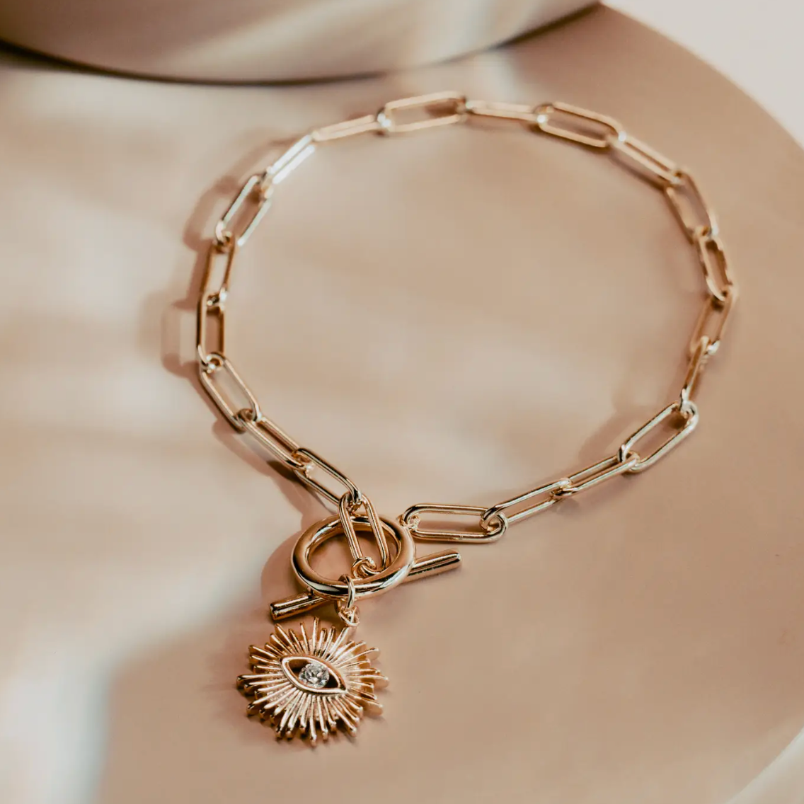The Golden Eye Bracelet blends sleek design with symbolic detail. Crafted in 14k gold plated over silver, it features a bold paperclip chain and a front toggle clasp accented by a radiant sunburst charm set with a clear crystal eye. Polished and versatile, it brings a touch of strength and modern refinement to any style.