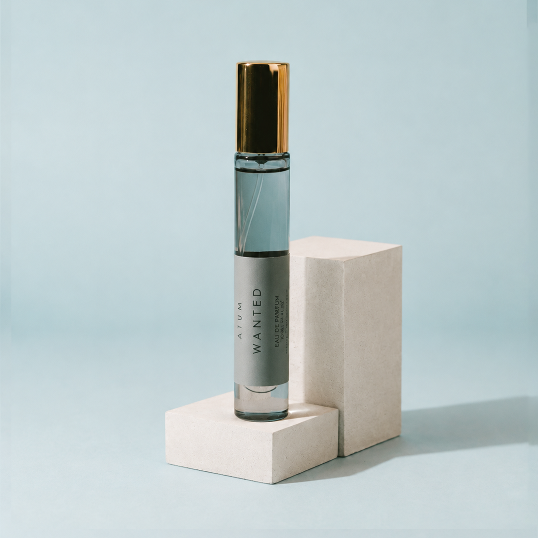A clear cylindrical perfume roller bottle with a metallic gold cap and a gray label reading 'ATOM WANTED', displayed on two stacked light beige blocks against a pale blue background.