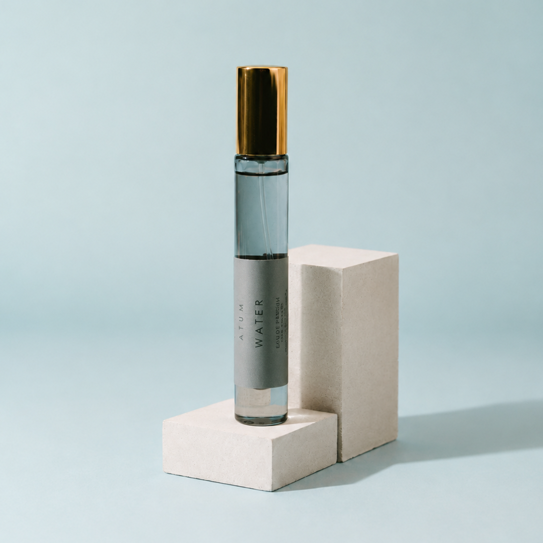 A clear glass perfume roller bottle with a metallic gold cap and a gray label reading 'A TUM WATER', standing on two stacked beige blocks against a light blue background.