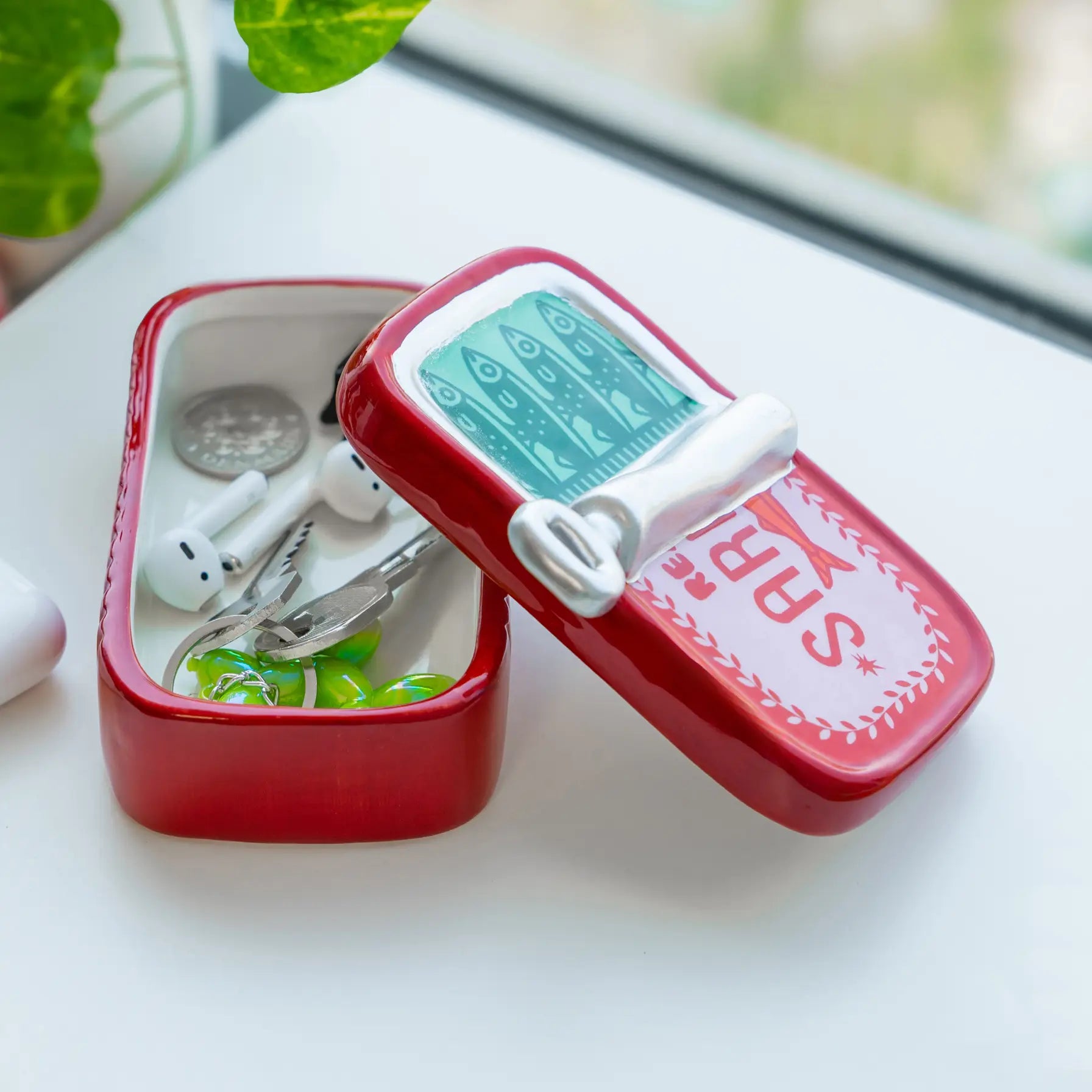 A small red metal first-aid kit tin with its lid propped open, showing contents like earbuds, a coin, and small green accessories. The lid has a printed medical-style face with the letters 'SRB' and a white clasp.