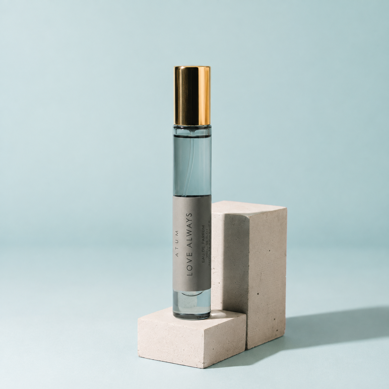 A clear glass roll-on perfume bottle with a metallic gold cap and a gray label reading 'ATUM LOVE ALWAYS', displayed on a light beige concrete block against a soft light blue background.