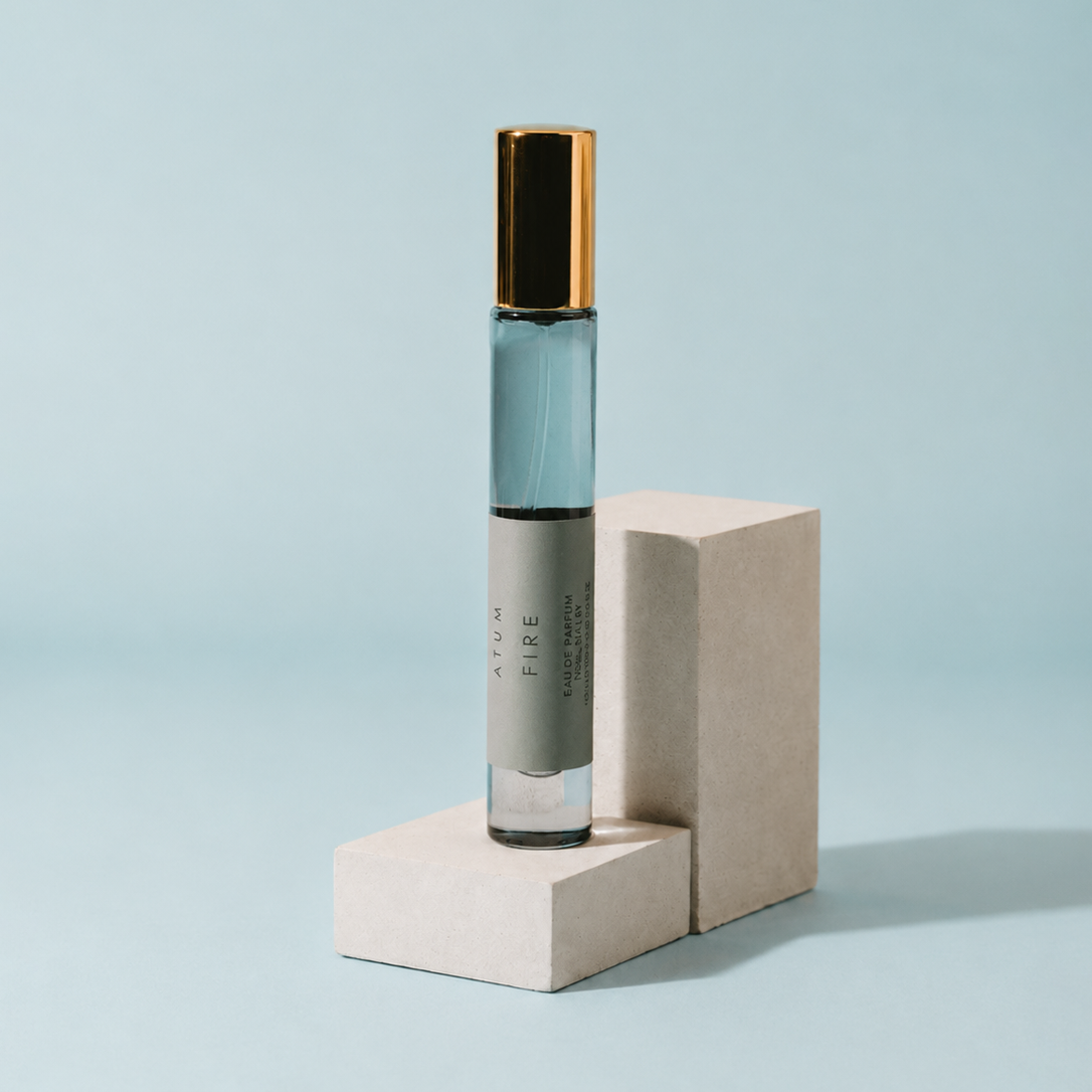 A clear glass roller bottle with a black cap and gold-toned collar, filled with pale blue liquid, standing on a light beige geometric display block against a soft light blue background.
