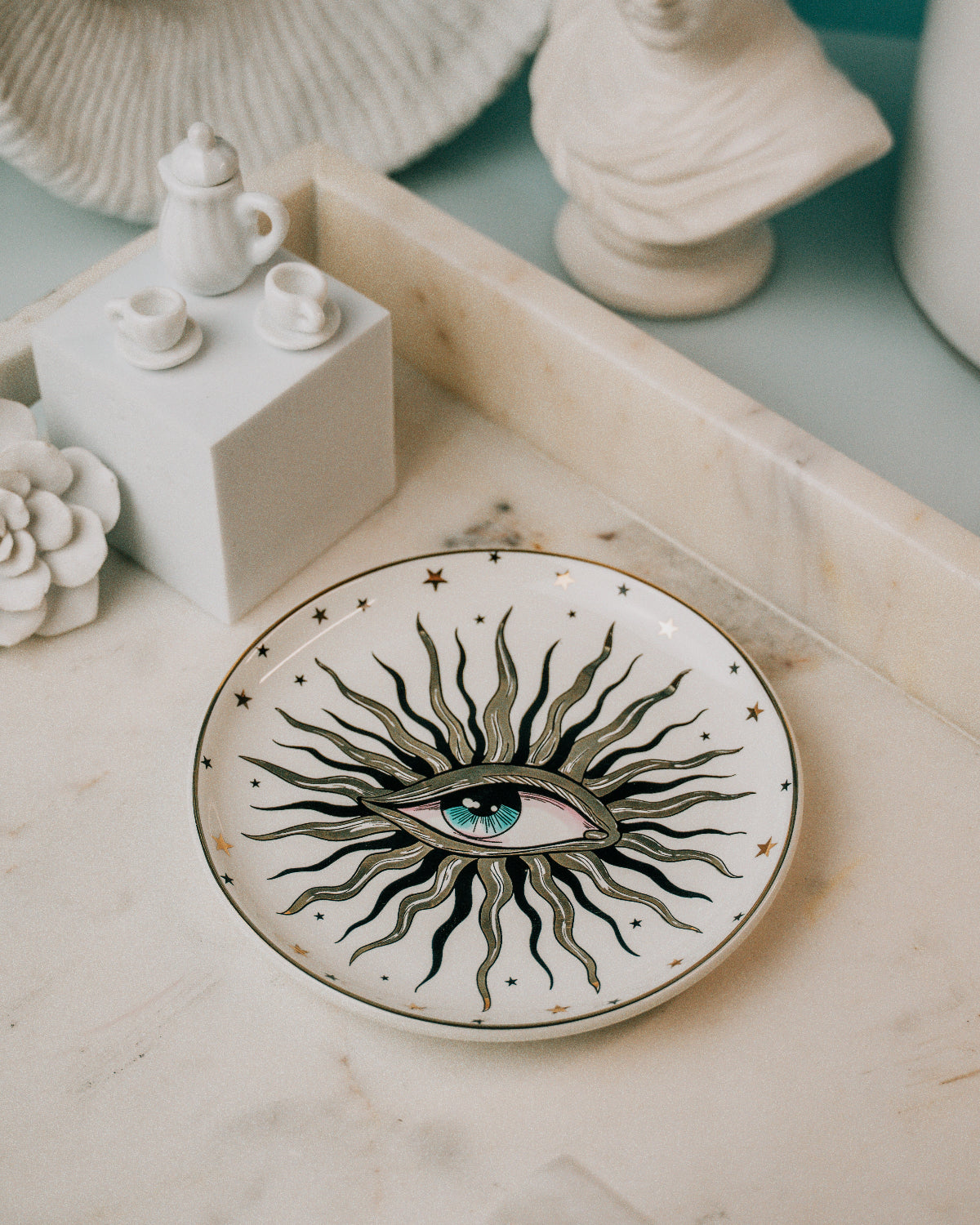 A round white ceramic plate with a hand-painted black and gray sunburst design radiating from a blue and green eye, decorated with small gold stars, displayed on a marble tray.