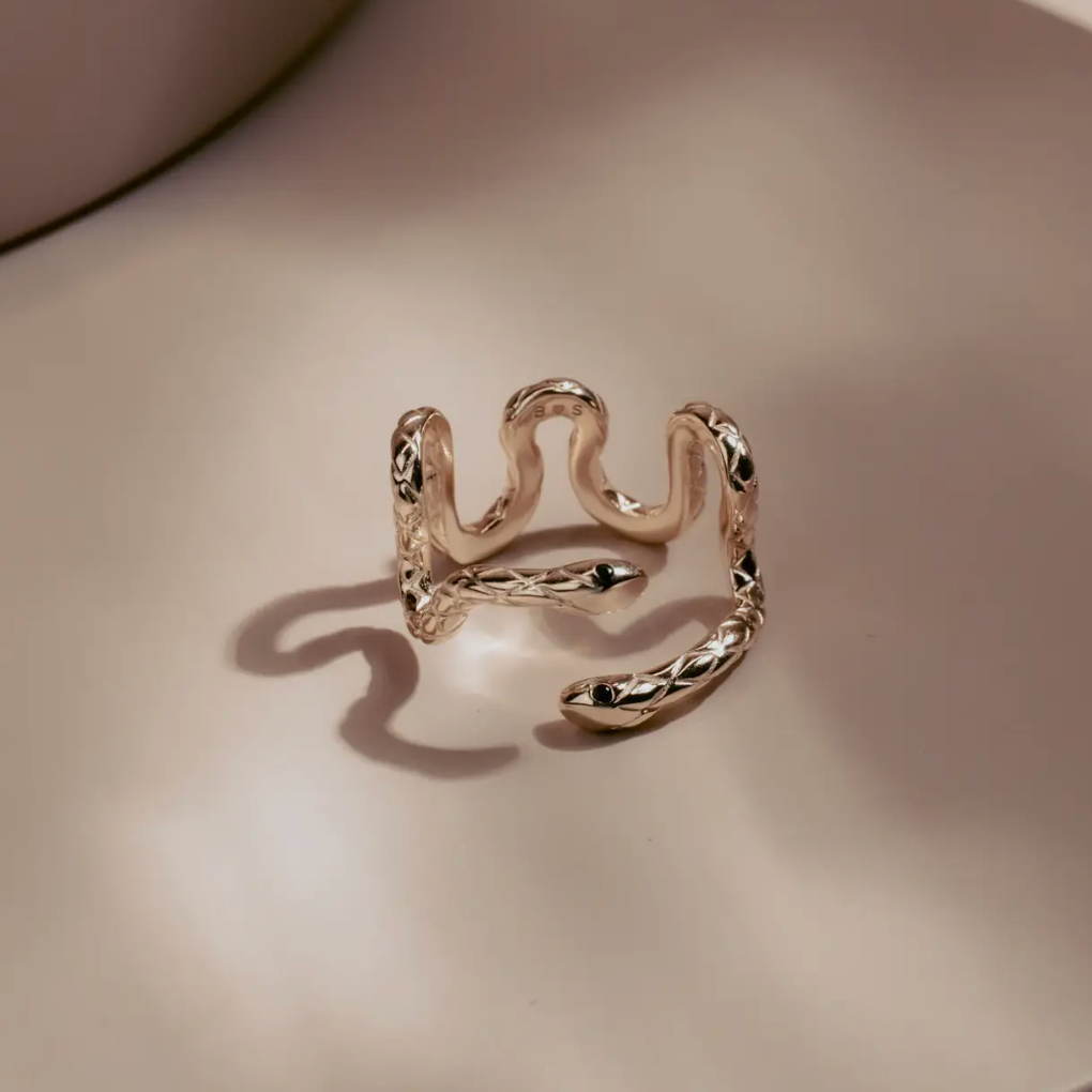 Sleek and hypnotic, the Serpent Coil Ring embodies power and transformation. Crafted in gleaming 14k gold plate over sterling silver, the coiled design wraps gracefully around the finger, ending in twin sculpted snake heads with dark alluring eyes. Adjustable for a perfect fit.