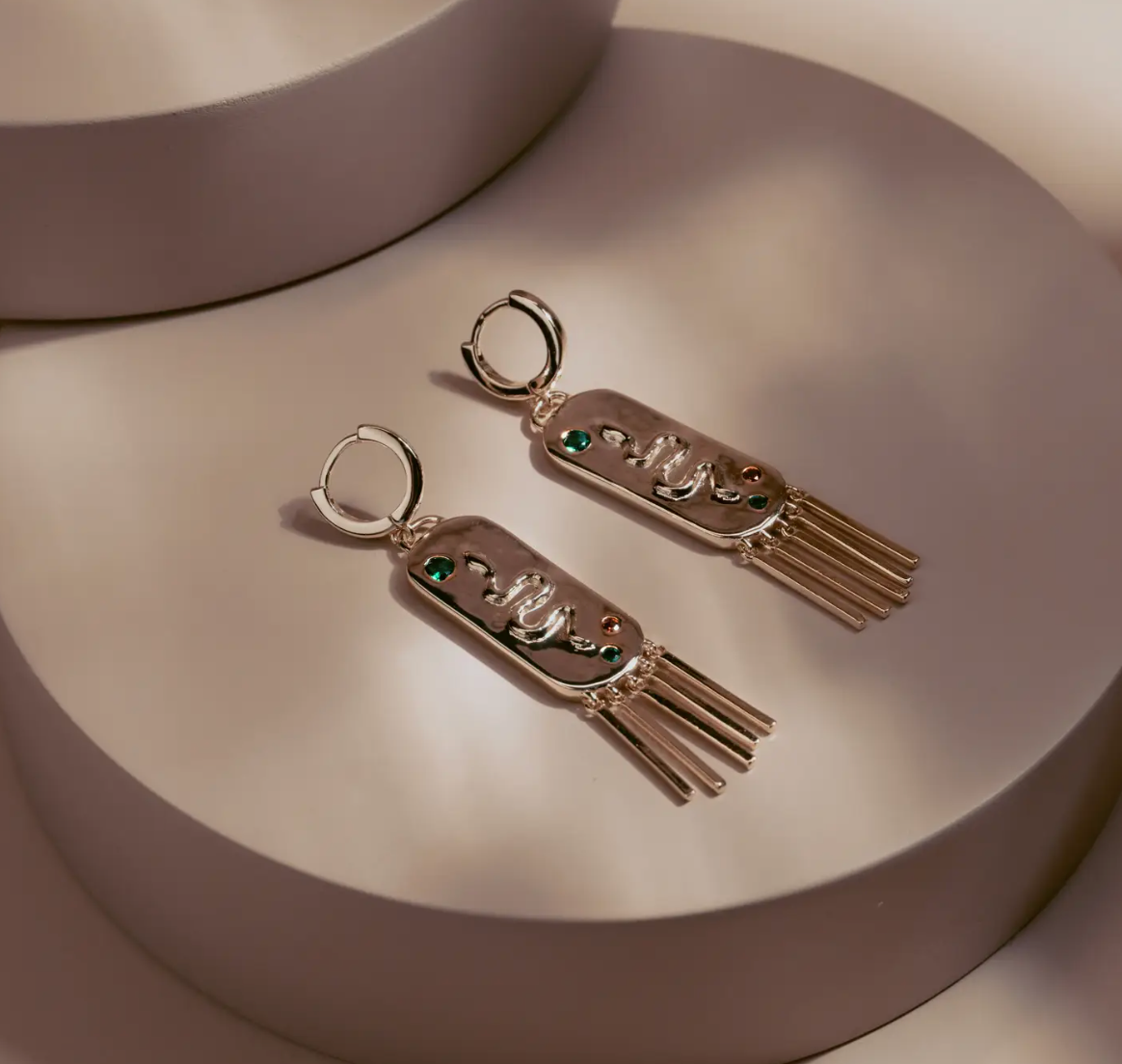 The Serpent Fringe Earrings fuse bold design with fluid movement. Crafted in 14k gold plated over silver, each piece features a raised serpent motif accented with deep green and red stones. The rectangular base flows into fine golden fringe, creating a striking play of texture and light. Confident and sculptural, they bring a touch of power and sophistication to any look.