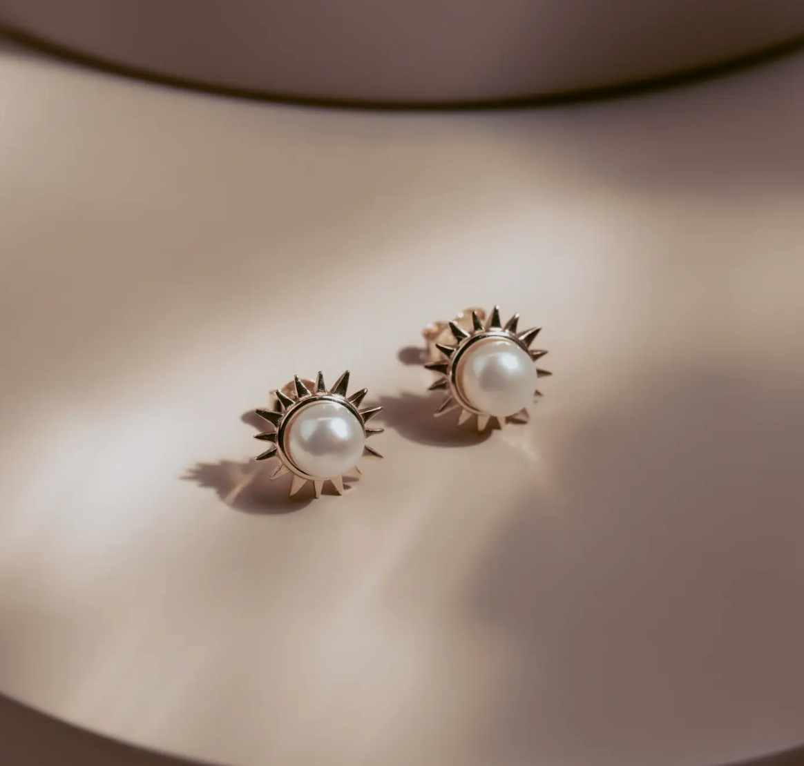 The Solar Pearl Studs capture the warmth of the sun in a timeless design. Each luminous freshwater pearl is framed by a burst of golden rays, crafted in 14k gold plate over sterling silver. Elegant yet bold, they bring effortless sophistication to any look—perfect for day or night.