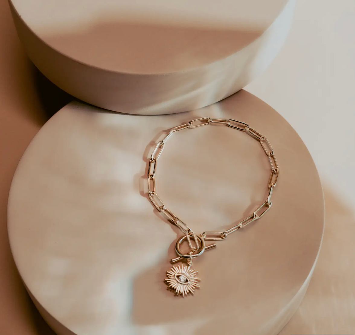 The Golden Eye Bracelet blends sleek design with symbolic detail. Crafted in 14k gold plated over silver, it features a bold paperclip chain and a front toggle clasp accented by a radiant sunburst charm set with a clear crystal eye. Polished and versatile, it brings a touch of strength and modern refinement to any style.