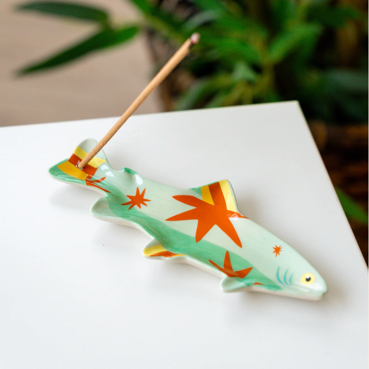 A ceramic fish shaped incense holder with orange starfish patterns and a wooden incense stick, displayed on a white surface with a blurred green plant in the background.