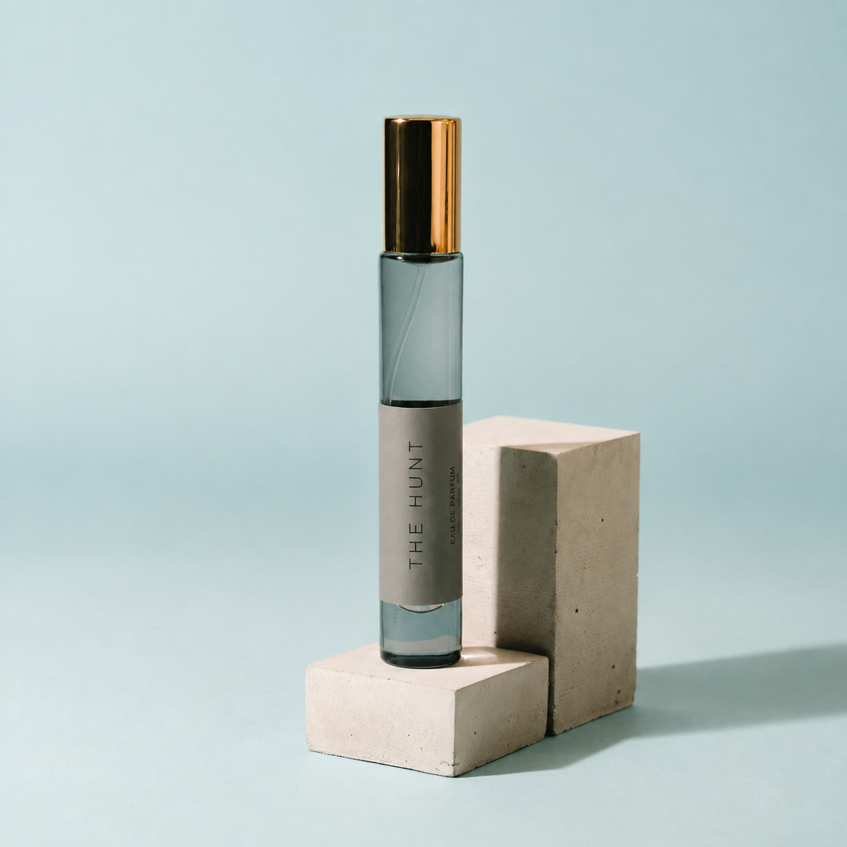 A clear glass roller bottle with a metallic gold cap and a gray label reading 'THE HUNT', displayed on two light beige concrete blocks against a soft light blue background.
