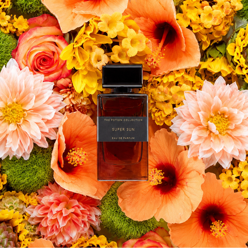 A rectangular glass perfume bottle labeled 'THE POTION COLLECTION SUPER SUN EAU DE PARFUM' sits centered on a dense floral arrangement of orange hibiscus, peach dahlias, yellow flowers, and green foliage.